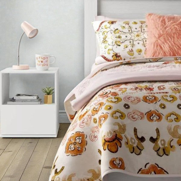 Bedroom with patterned white and orange bedding, pink fluffy pillow, white nightstand, and pink lamp