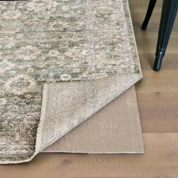 Close-up of a beige and green patterned rug corner lifted to show a non-slip rug pad underneath on hardwood floor