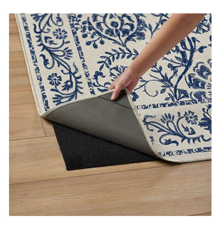 Hand lifting corner of blue and white floral area rug showing black non-slip rug pad on wooden floor