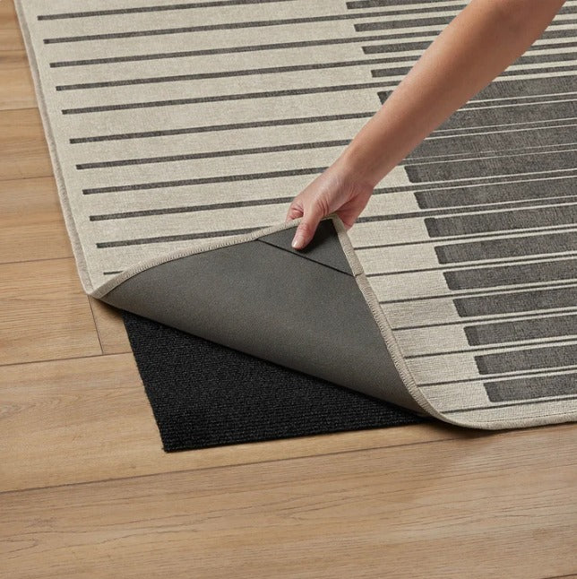 Hand lifting the corner of a beige and gray striped area rug revealing a black rug pad underneath on wooden floor